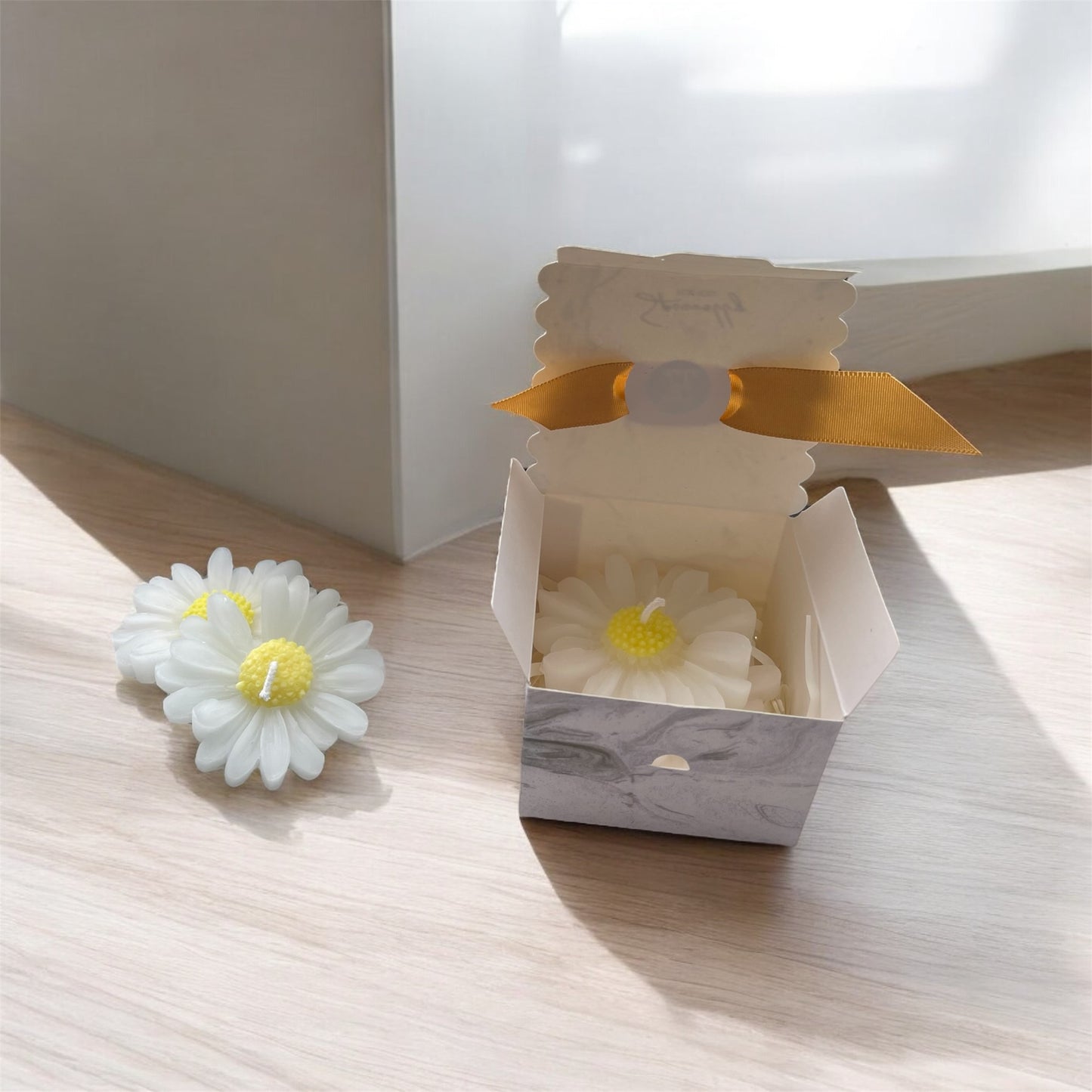 White daisy-shaped candle with yellow center on a wooden surface, next to an open box containing another similar candle.